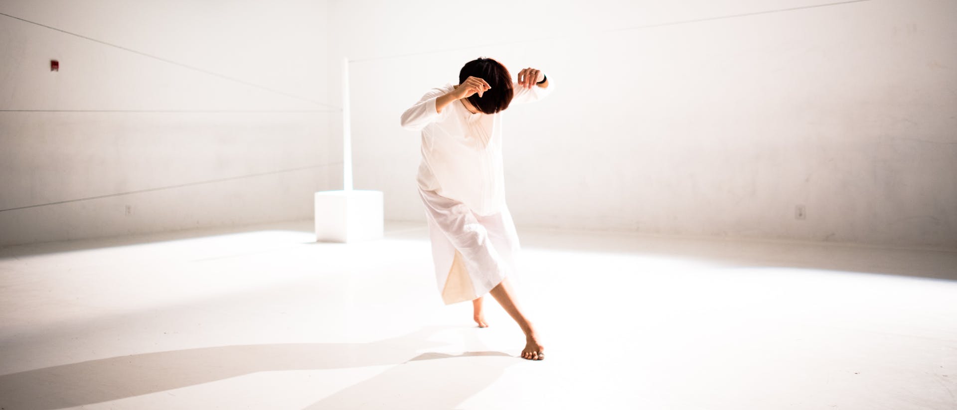 Hsiao-Jou Tang is centered, dancing in a brightly lit, bare white room. She is bent forward as she steps, head tilted down, with her elbows raised by the sides of her head. She is wearing a long sleeved, loose white dress.