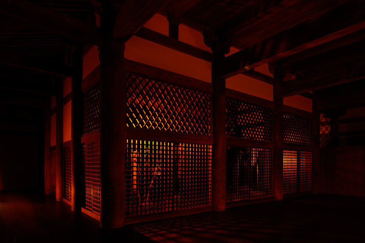 Within a dark, red lit room, a space is enclosed by walls with grid-like openings. Thick wooden columns stand at the corners of these walls and thick wooden bars fill the ceiling. 