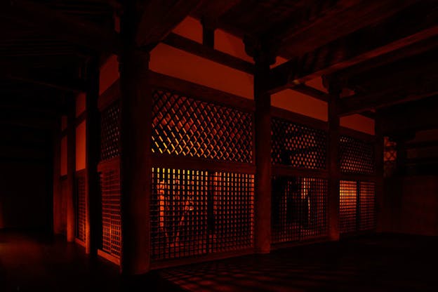 Within a dark, red lit room, a space is enclosed by walls with grid-like openings. Thick wooden columns stand at the corners of these walls and thick wooden bars fill the ceiling. 