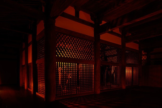 Within a dark, red lit room, a space is enclosed by walls with grid-like openings. Thick wooden columns stand at the corners of these walls and thick wooden bars fill the ceiling. 