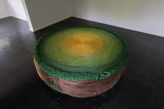 A round, squat sculpture made from braided synthetic hair. The top portion is a large disc that is yellow at the center, and transitions outward to dark green with thick green braids lining the border of the disc. The base rounded towards the bottom and is constructed of light brown synthetic braids.