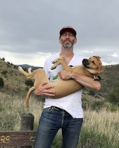 Image of Richard Maxwell, standing, holding a dog in his arms in front of a mountainous backdrop.