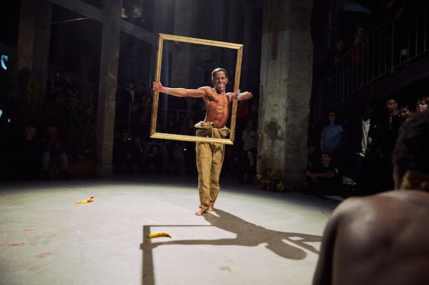 A performer is centered, standing in a concrete performance space holding a large empty picture frame. They are barefoot and wearing tan coveralls with the top lowered and the arms tied around their bare waist. They wear a smiling Obama mask. 