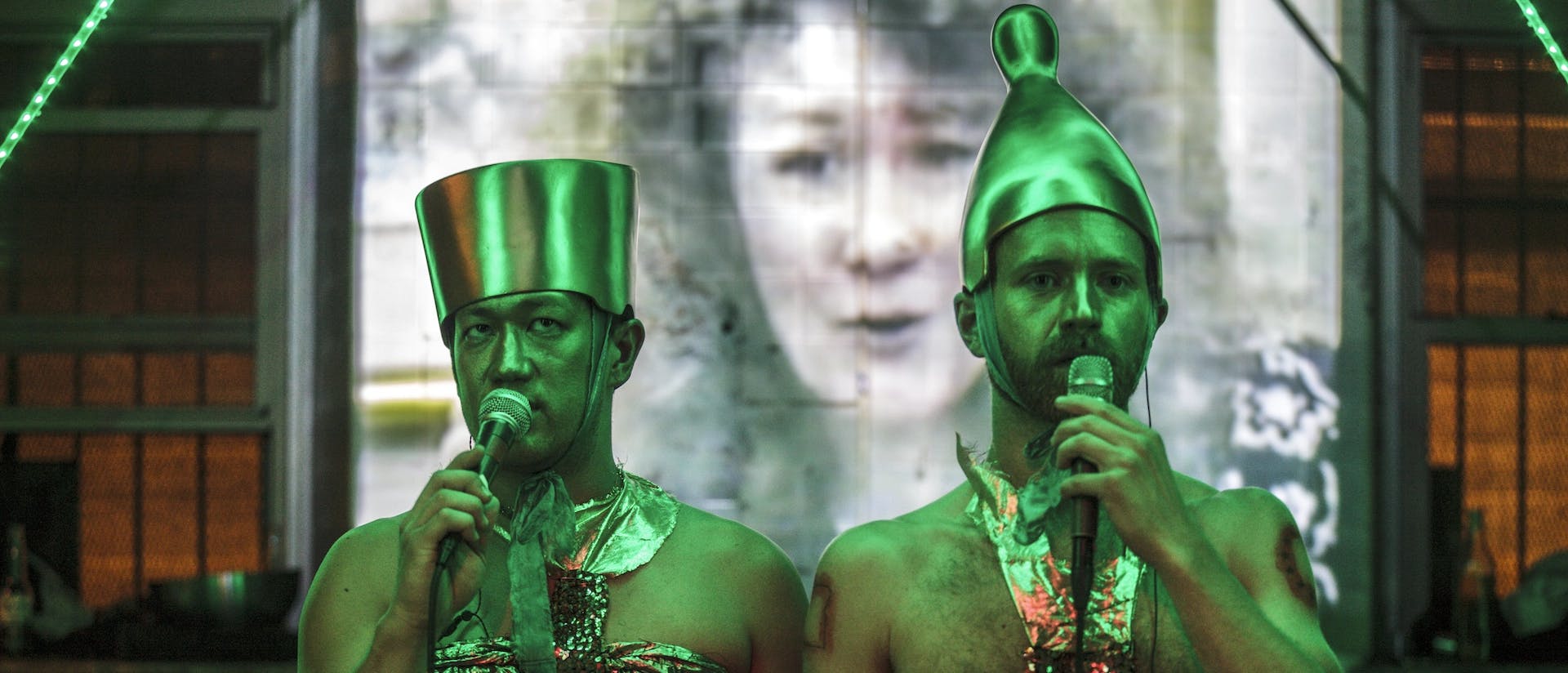 Tei Blow and Sean McElroy stand side by side, vocalizing into microphones and bathed in green light. They stand in sharp focus in front of a projection screen where there is a woman's face displayed out of focus. Both are wearing large golden headpieces and golden halter tops made of metallic, sparkly fabric. Blow's headpiece is flat-topped where McElroy's is tapered and cone-shaped with a rounded tip. 