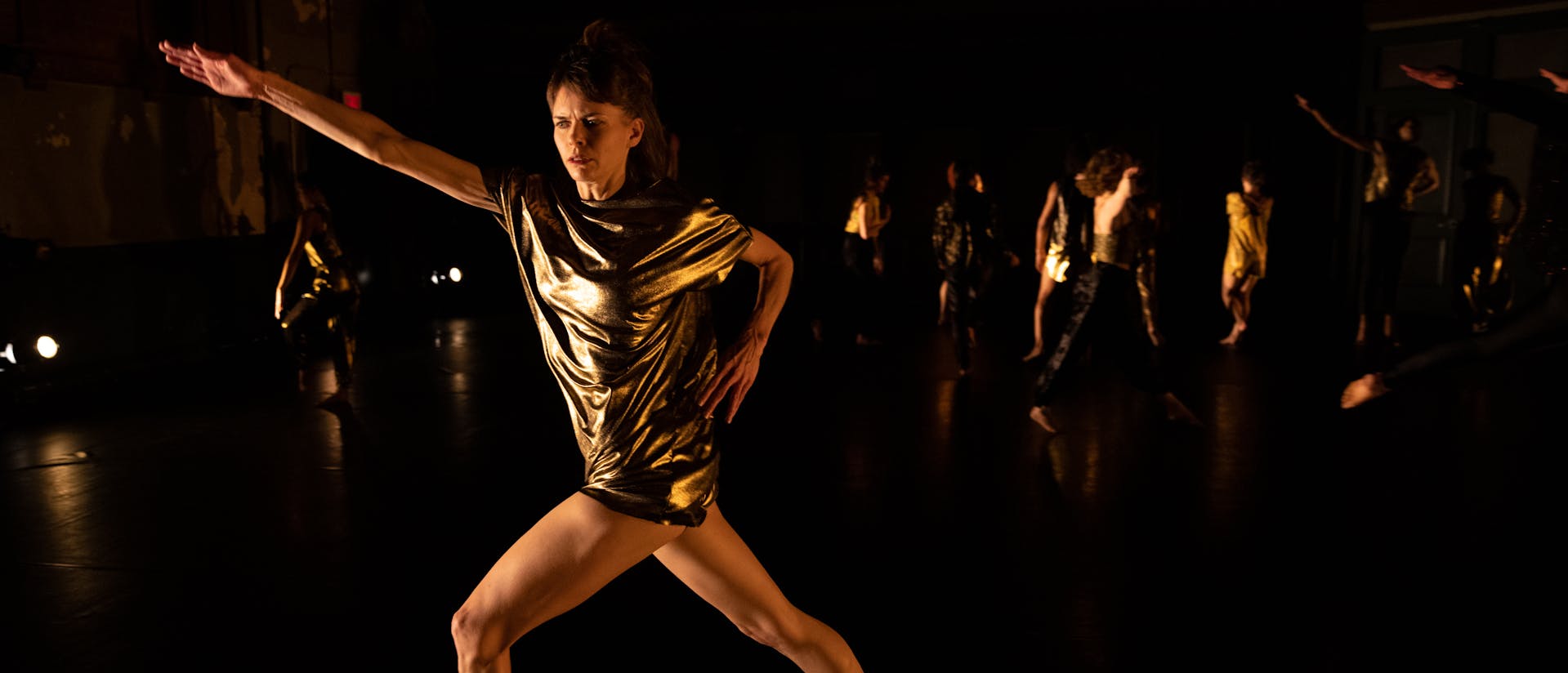 Joanna Kotze is centered as she performs on a dark stage with minimal yellow lighting, some dancers are visible in the far background behind her. She looks towards the left with concentration as she lunges forward on her left leg, hips pointing to the left. Her torso twists as her shoulders angle towards the camera. Her left arm is bent as her hand rests on her left hip, and her right arm is extended and reaches out and up to the left where she lunges, hand outstretched with her palm facing down. She wears a gold metallic shortsleeved tunic.