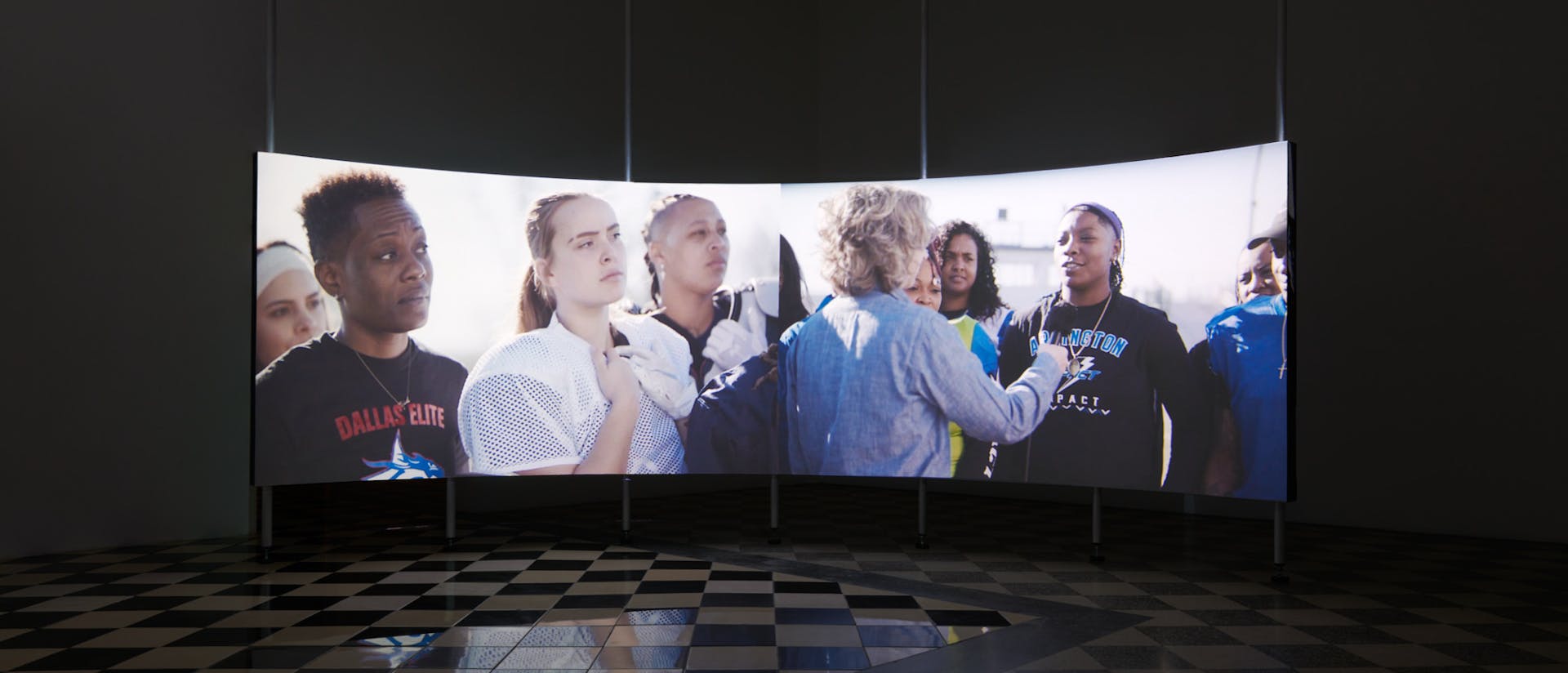 A dark gallery space with checkered stone floors and a large, concave projection screen curved to fit the shape of the room. On the projection screen, two video frames are displayed. On the left half of the screen is an image of four women athletes turned to listen to an offscreen speaker. One is wearing a crewneck that says, 