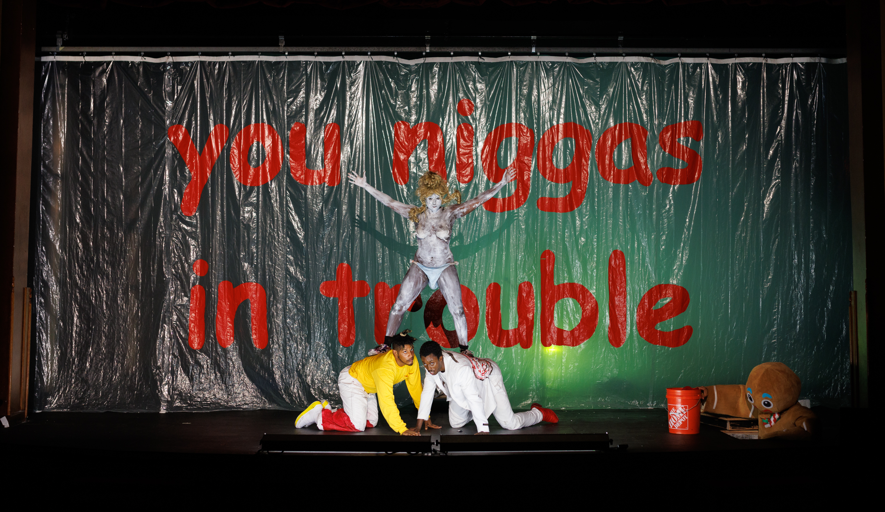 Three performers are centered at the front of a stage, lit with stark white light from below against a green backdrop. Behind them is a clear plastic curtain that reads in large red lowercase text, 
