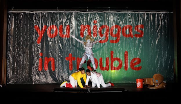 Three performers are centered at the front of a stage, lit with stark white light from below against a green backdrop. Behind them is a clear plastic curtain that reads in large red lowercase text, 