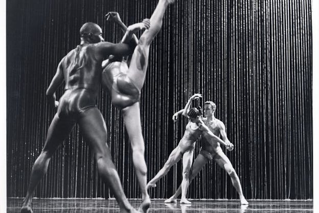 A black and white photograph of two pairs of performers dancing in front of a rain curtain. The first pair in the front has their backs facing the camera. One of them raises their right leg straight up while the other one holds their arms. The second pair in the back faces directly to the camera. One of them slightly raises their right leg while the other one holds their hands. The stage is set against a dark backdrop.