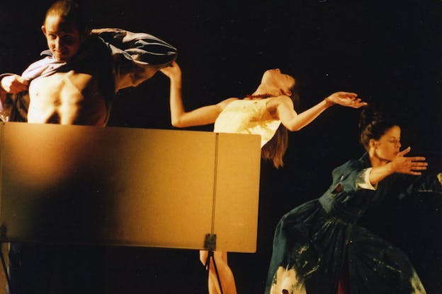 Three performers are in a dark space with a black background and a small tan panel in front of them. The performer on the left puffs their chest out and pulls off their shirt. The performer in the middle wears a white dress and leans their head back and holds their arms up by their shoulders. The performer on the right wears a blue dress and bends down, holding their arms in front of their chest. 