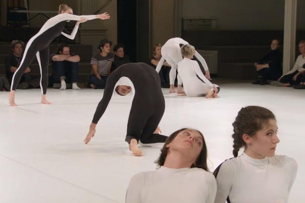 Performers dressed in full body leotards, white in the front and black at the back, lay down in various poses on a white rectangle floorboard. The two closest two the viewer sit next to each other looking sideways. 