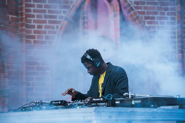 A DJ dressed in a yellow shirt and black dress jacket stands behind a DJ controller while smoke engulfs them. 