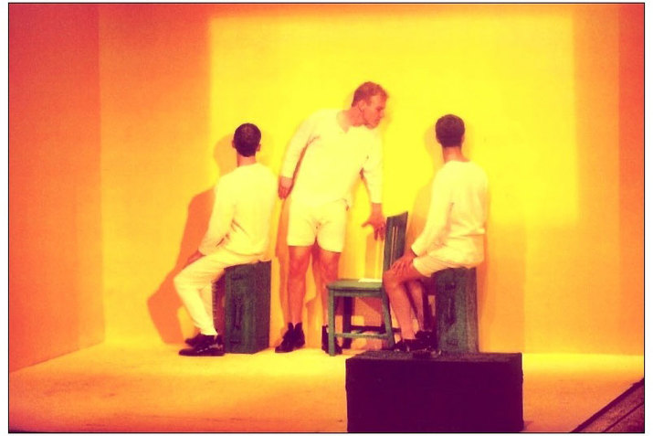 Three performers wearing all white stand in an orange hued bare cube. Two performers sit on dark wooden crates with their bodies angled towards the left wall and their heads turned  towards the back wall. A third performer stands between them and bends over to face the performer on the right. 