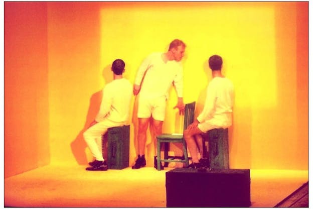 Three performers wearing all white stand in an orange hued bare cube. Two performers sit on dark wooden crates with their bodies angled towards the left wall and their heads turned  towards the back wall. A third performer stands between them and bends over to face the performer on the right. 
