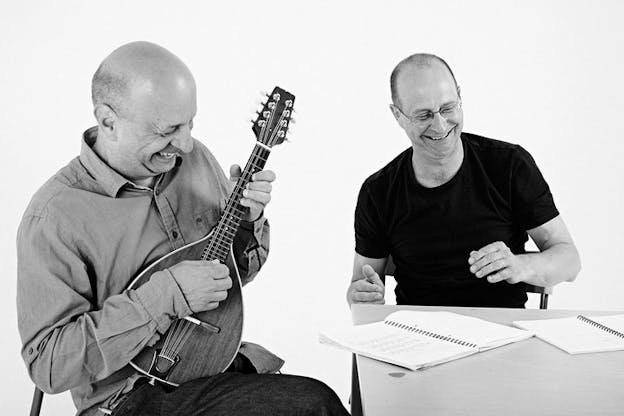 A black and white photograph of two people sitting at a table in front of a white background. On the left a person sits and plays the mandolin. The person on the right looks down at opened notebooks. Both people appear to be laughing.