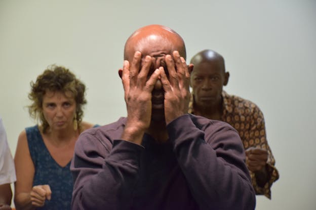 A figure en face the viewer dressed in a maroon shirt covers their eyes and face with their hands. Two people stand behind them watching with their hands in raised fists.