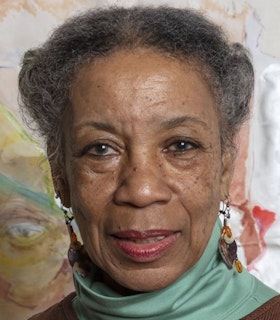 Suzanne Jackson looks directly into the camera, standing before an abstract painted background of mostly cream and yellow hues. She is wearing a mint green turtleneck under a brown sweater, and dangling earrings. 