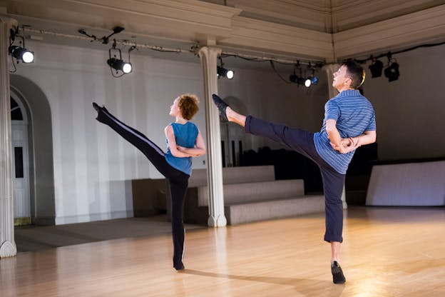 Side-angle photograph of two aligned dancers raising their left leg, standing on relevé and looking outwards. 