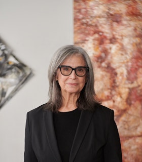 Judith Geichman looks directly at the camera with a slight smile. She stands in front of two of her paintings wearing a black top, a black blazer, and glasses.