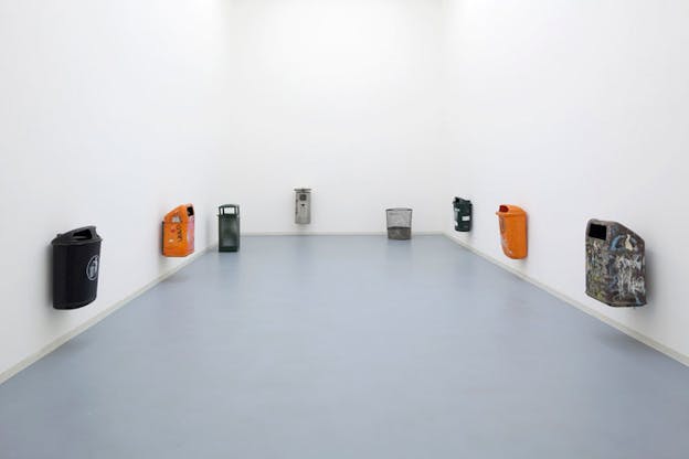 An installation image of eight trash cans in a white room. Most of the trash cans are hung on the wall but two rest directly on the floor. The cans are various colors including orange, grey, and black. Some of the cans are covered in graffiti while others have printed signage on them. 