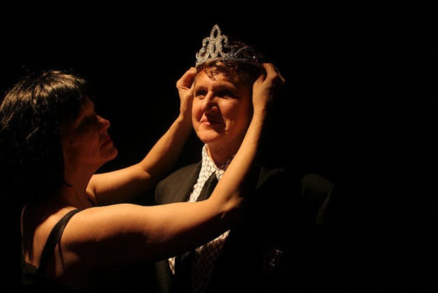 Peggy Shaw is in front of a black background, wearing a black suit, a black tie, and a black and white patterned shirt. Another person, with a black bob haircu places a plastic tiara on Peggy Shaw's head. This person is mostly hidden in shadows and appears focused while Peggy Shaw is bathed in warm light and has a proud and content expression.