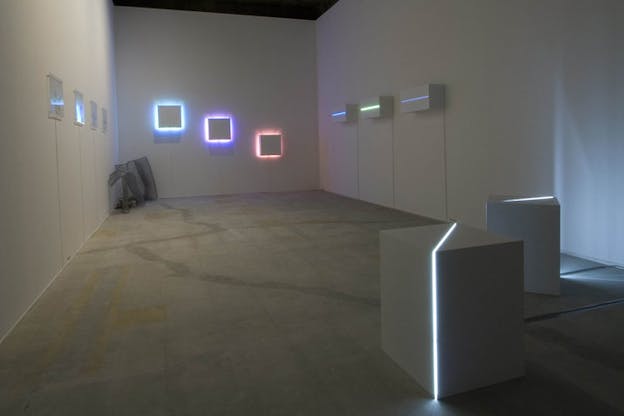 A minimally furnished gray space contains squares of different sizes on the wall. They are lighted in blue, purple and red colors.
