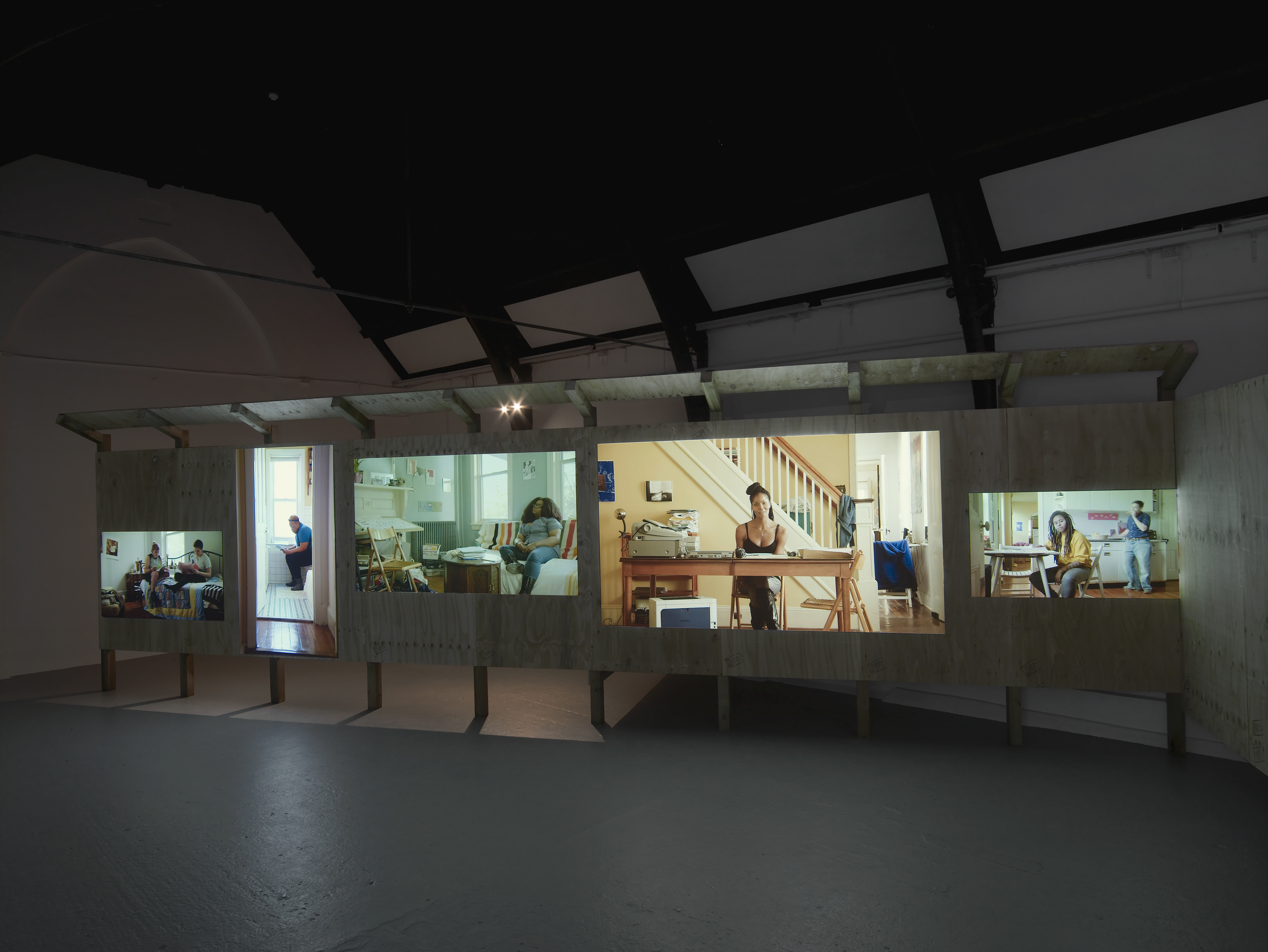 A dark gallery space with sloped ceilings holds a wooden panel board on which images of people sitting in their homes are projected.