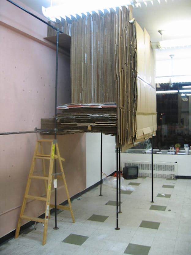 An installation image of a metal shelf filled with many large flattened cardboard boxes oriented vertically. On the upper left of the shelf, a smaller shelf was constructed high up by the ceiling and smaller bits of cardboard are stored there. In front of this shelf, there is a small wooden ladder. The walls of the room are pink and the floor if white with green tiles. In the far background, there is a large window which indicates that it is night time.