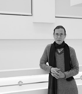Black and white portrait of Yvonne Rainer wearing thin round glasses, a black scarf, and a long grey sweater. The artist is leaning against a white wall with a protruding metal bar.