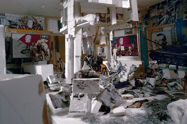 In a cluttered studio-setting cut-outs of faces, bodies, blurred objects, a tank, are juxtaposed with prints of a man's face resembling the generic 20th century dictator image. 