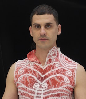 A portrait of Matt Savitsky in front of a black background. He wears a red shirt with white designs on it. He looks directly at the camera. 