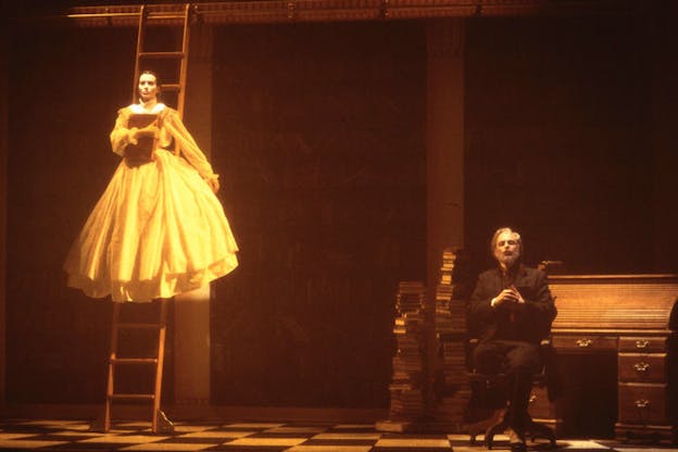 A woman in a yellow puffy dress stands on top of a ladder holding a book while a man to her right sits on a chair next to a pile of books. 