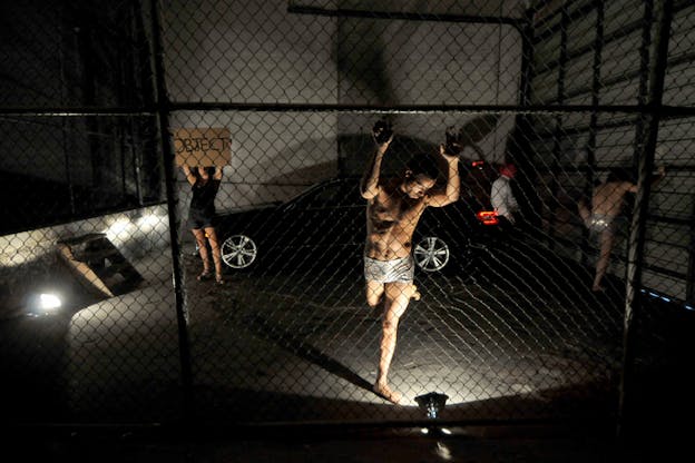 In a dimly-lit space enclosed by chain-link fence and gray walls, one performer wearing only silver shorts hangs onto the fence and raises one bent leg behind them. In the background beside a parked black car, one performer holds a cardboard sign written: 