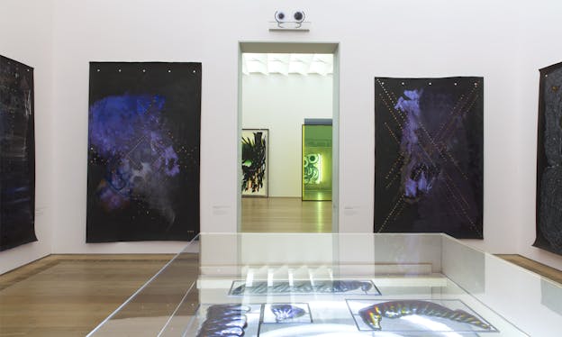 A white gallery room hosts two black cloth material canvases with blurs of purple and blue, small bronze circles align intersectionally. Between the two canvases a door frame to another room showing the half of a transparent green glass on the right and a canvas with trails of black, green and yellow creating leaves.