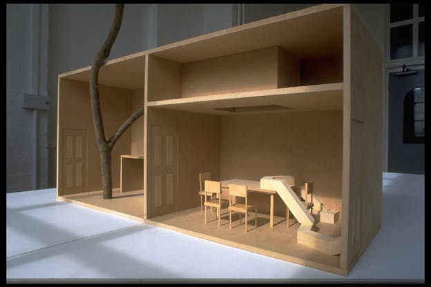 A wooden house model is staged on a white surface. Inside one room of the model a tree branch seems to grow.