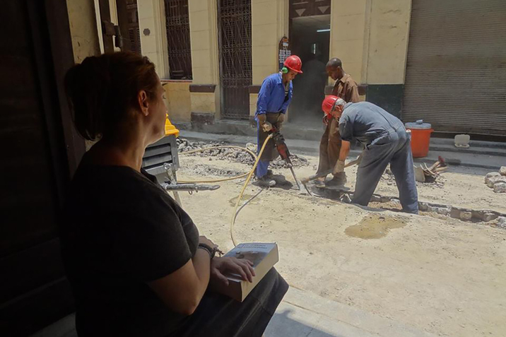 A figure sideways to the viewer sits with a book on the lap watching construction workers dressed in jean blue overalls and red safety hats drill on a dirt road.