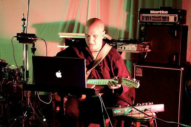 David First wears a loose dark shirt and plays a guitar in his lap. He looks intently at the screen of a laptop in front of him. Behind him, there is more musical equipment in front of a white sheet which appears to hang from the ceiling and reaches the ground. The scene is bathed in red and green light.