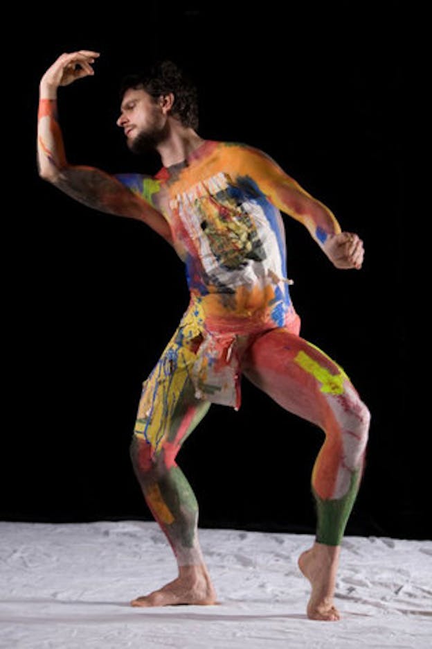 A dancer whose body is painted from neck to ankle in earthy oranges, reds, greens, and blues strikes a pose with one foot arched, bending their head towards their arm curved in a crescent-shape. The background is black and the floor is covered in white cloth. 