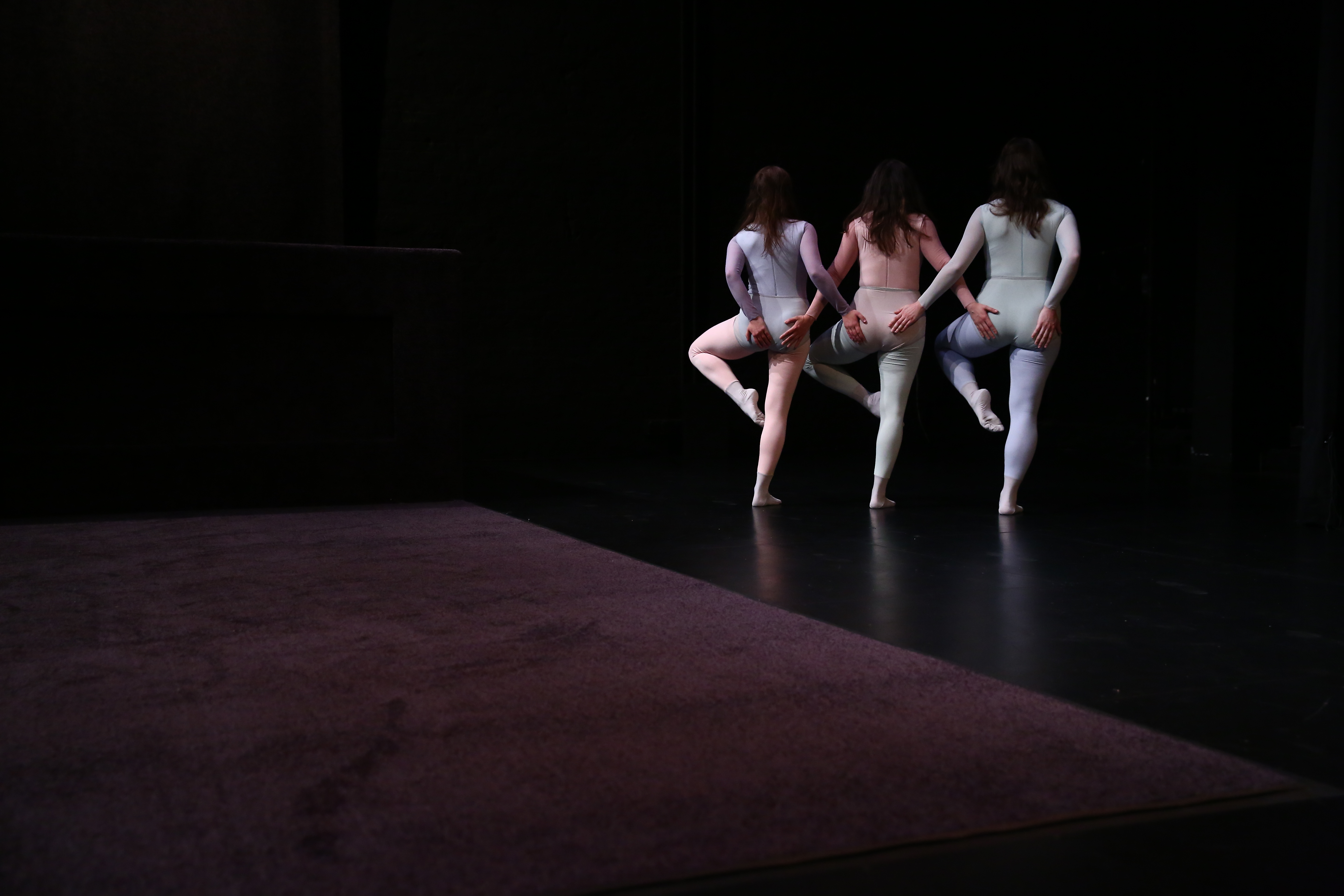 Three performers with their backs towards the viewer with entwined arms touching each others derrières have their left leg angled upwards to their side.