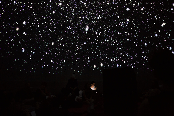 A figure sits in darkness surrounded by specs of white stars projected on the black wall.