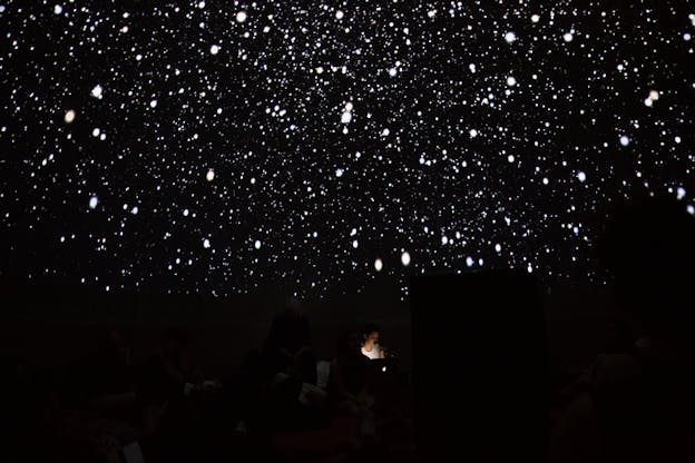 A figure sits in darkness surrounded by specs of white stars projected on the black wall.