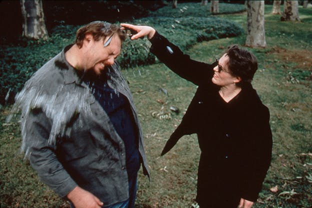 A photograph of Matin Kersels being smacked by a person wearing all black with short brown hair and sunglasses. Kersels winces and water drops are seen splaying off of him. He wears a grey jacket and a navy shirt. They stand on grass in front of an area with bushes and trees. 