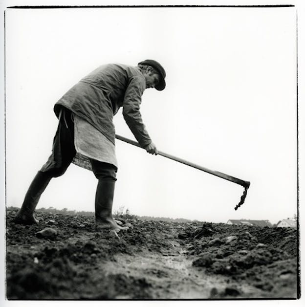 Black and white photograph showing a person  a person's profile working field, dressed in a coat, long shirt, plastic boots and a hat holding a hoe.   