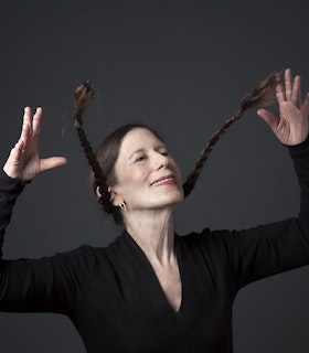 A portrait of Meredith Monk dressed in a black V-neck shirt, small hoop earrings and red lipstick throws with her hands her brown pigtails forward while her head is titled back smiling.