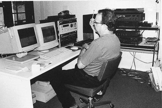 A black and white photograph of Carl Stone sitting at a desk and looking at two computer desktops. To his left, there are several sound machines. Stone wears a gray t-shirt, dark jeans, and holds a pencil in his hand as if lost in thought. 