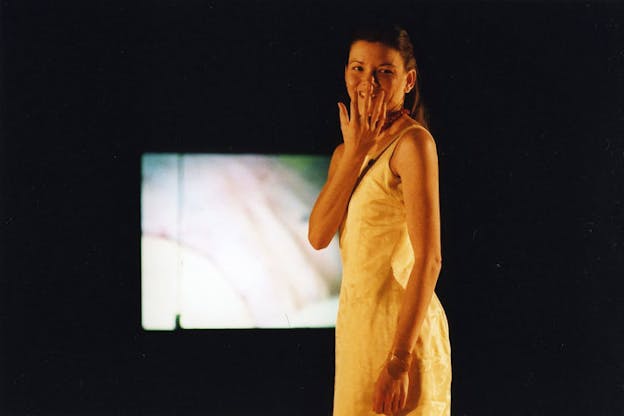 A performer wearing a white dress faces the camera and smiles, putting a hand in front of their mouth. The background is all black aside from a blurry projection in the distant background.