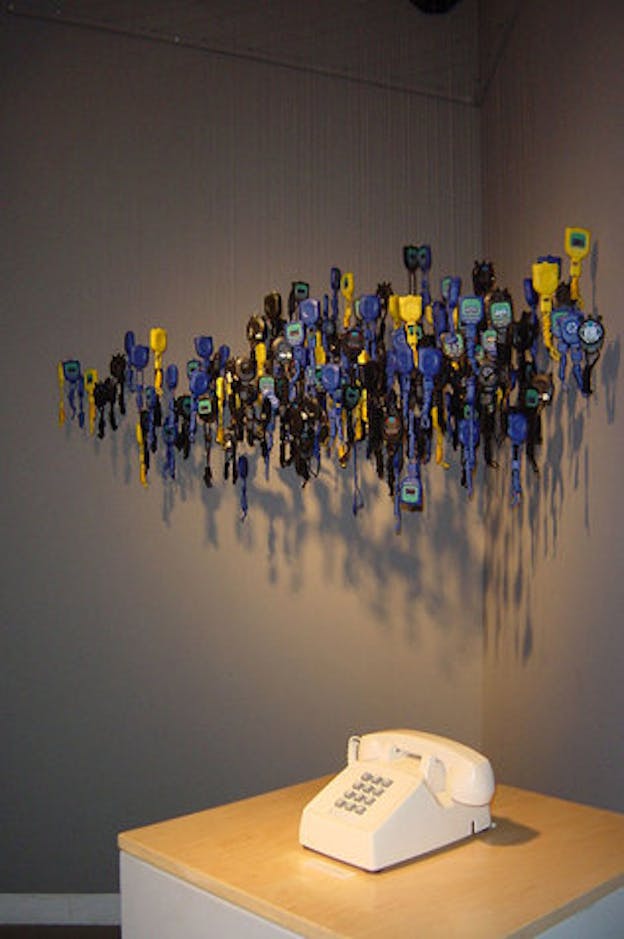 An installation image of a white rotary telephone on a wooden table. Above it, numerous yellow, black, and blue mechanical objects are suspended in a cluster by translucent wire. 