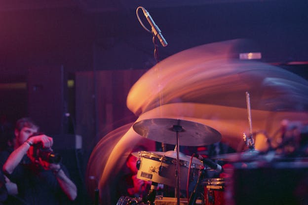 A motion blur photograph of drums and a microphone lighted by orange pink lights, smooth lines sit above them.