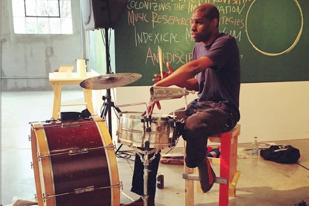 A person seated on a colorful wooden high bench playing percussion instruments. Around them on the floor are scattered water bottles and black bags, while behind them a green eraser board has a cirle drawn on it and next to it words and phrases like 