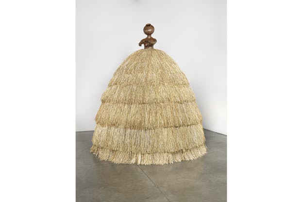Image of hay straws forming a silhouette similar to that of a hoop skirt. On top of it a wooden brown statue with a round head an no features.